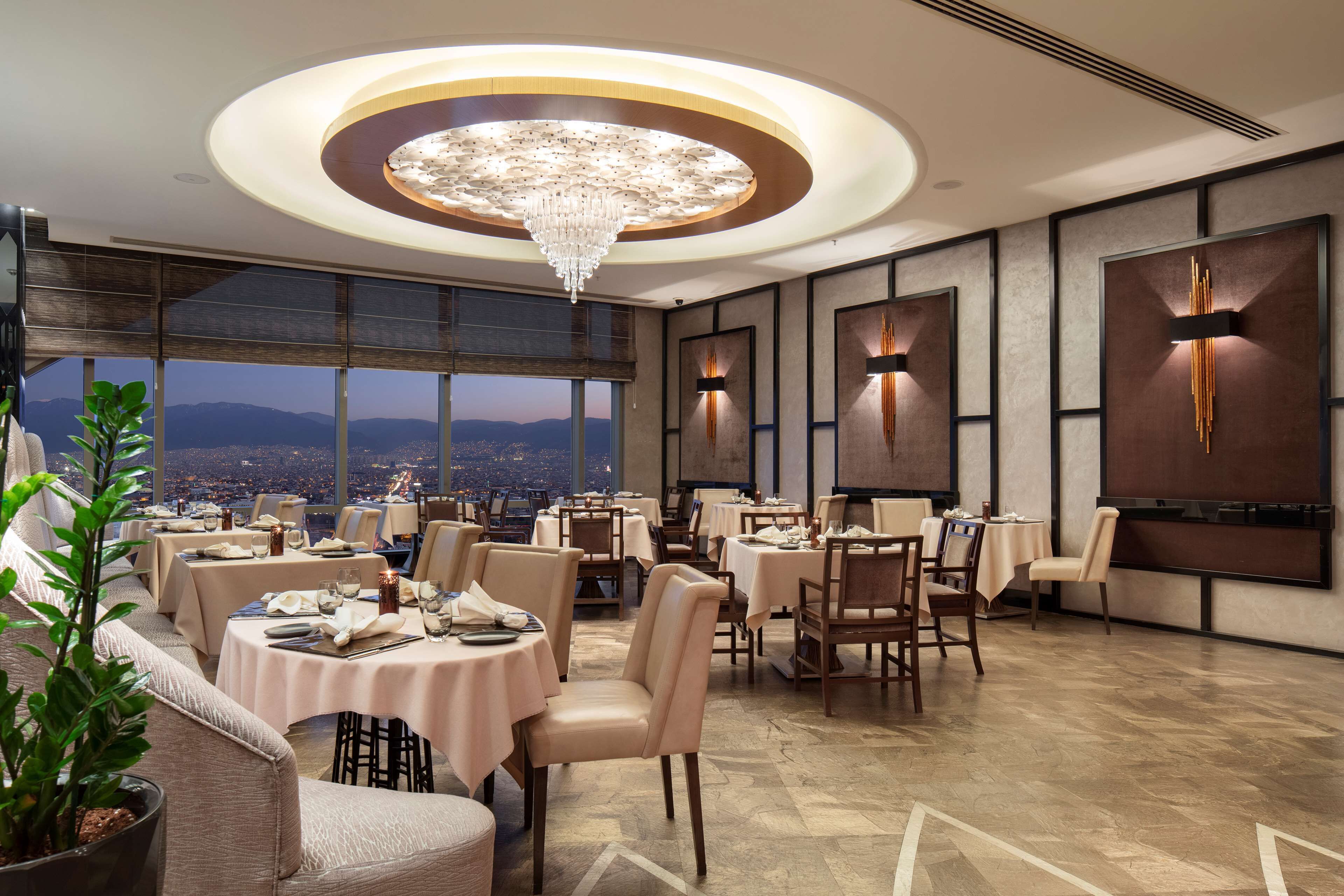 Un restaurante moderno con mesas redondas, sillas elegantes y una lámpara de araña grande. Ofrece vistas a la ciudad a través de amplias ventanas y paneles decorativos en las paredes.