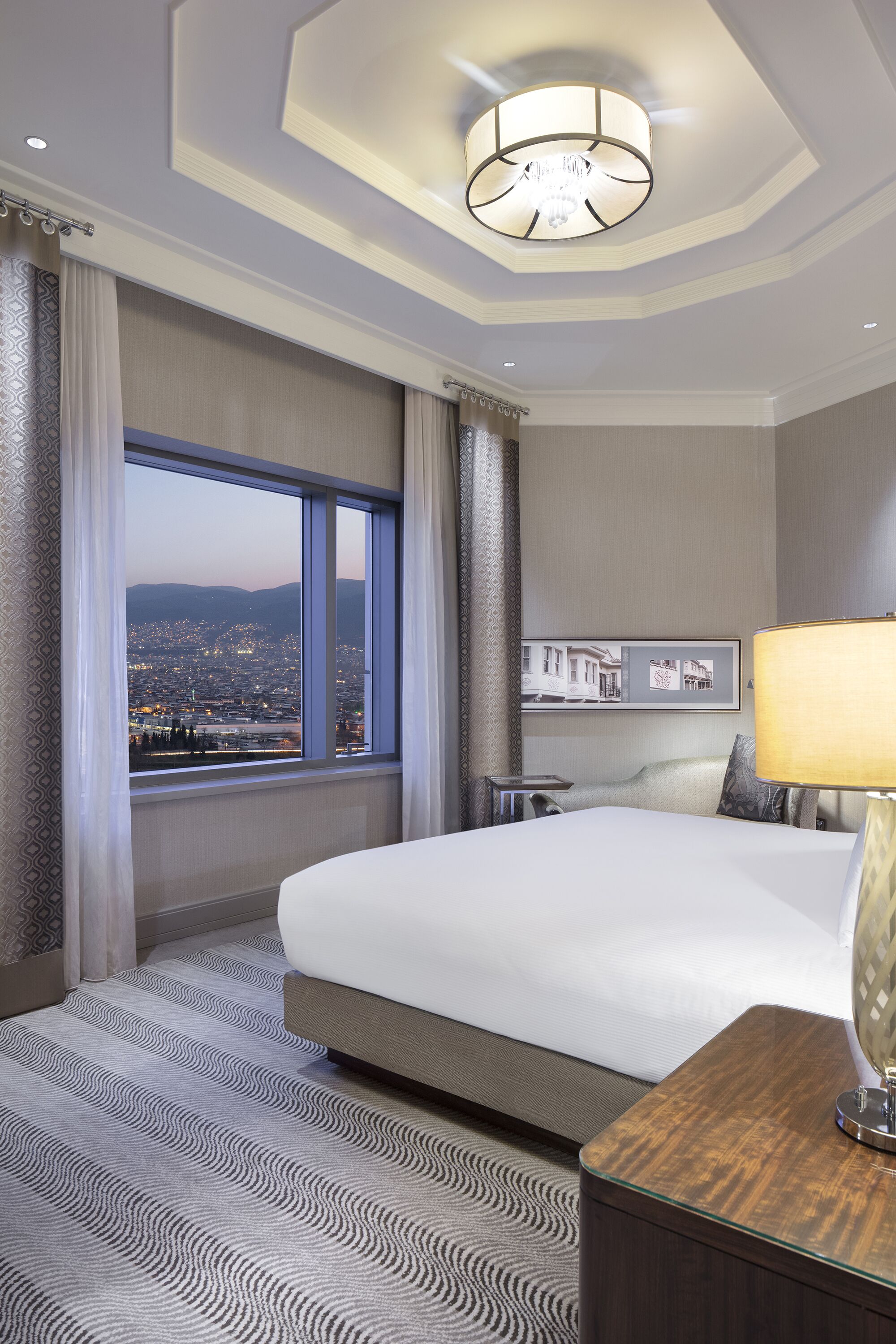 A hotel room with a large bed, a view of the cityscape through a large window, a chandelier, and a nightstand with a lamp.