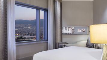 A hotel room with a large bed, a view of the cityscape through a large window, a chandelier, and a nightstand with a lamp.