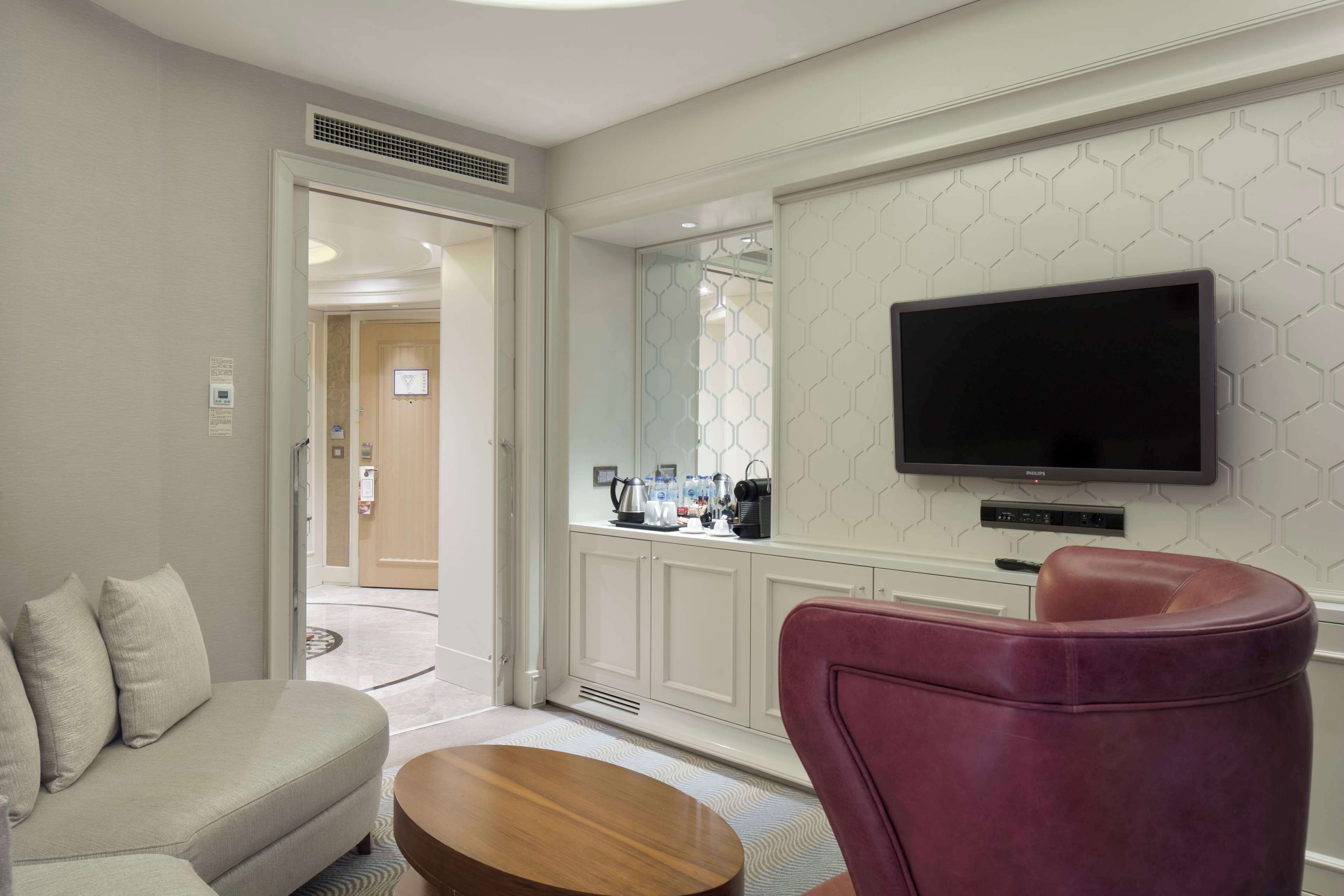 Una habitación de hotel moderna con sofá, una silla roja, una mesa de centro de madera, un televisor de pantalla plana montado en la pared y una cocina visible a través de un panel de vidrio.