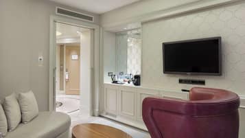 Una habitación de hotel moderna con sofá, una silla roja, una mesa de centro de madera, un televisor de pantalla plana montado en la pared y una cocina visible a través de un panel de vidrio.