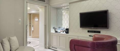 A modern hotel room with a sofa, a red chair, a wooden coffee table, a flat-screen TV mounted on the wall, and a kitchen area visible through a glass partition.