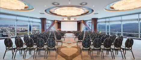 A conference room with rows of chairs facing a large screen, large windows offering a city view, and modern chandeliers.