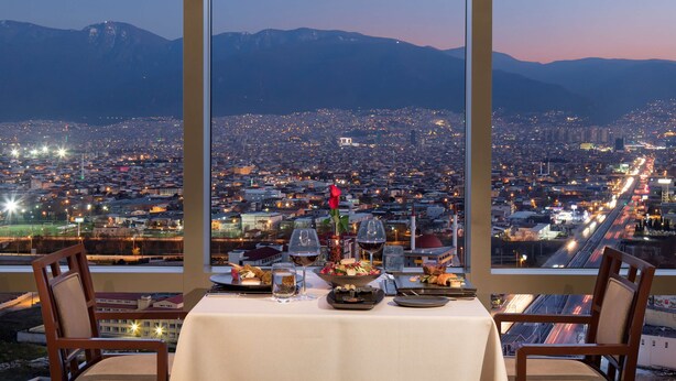 Une table à manger avec vue sur la ville, deux chaises et une table dressée comprenant des verres à vin et un vase à fleurs.