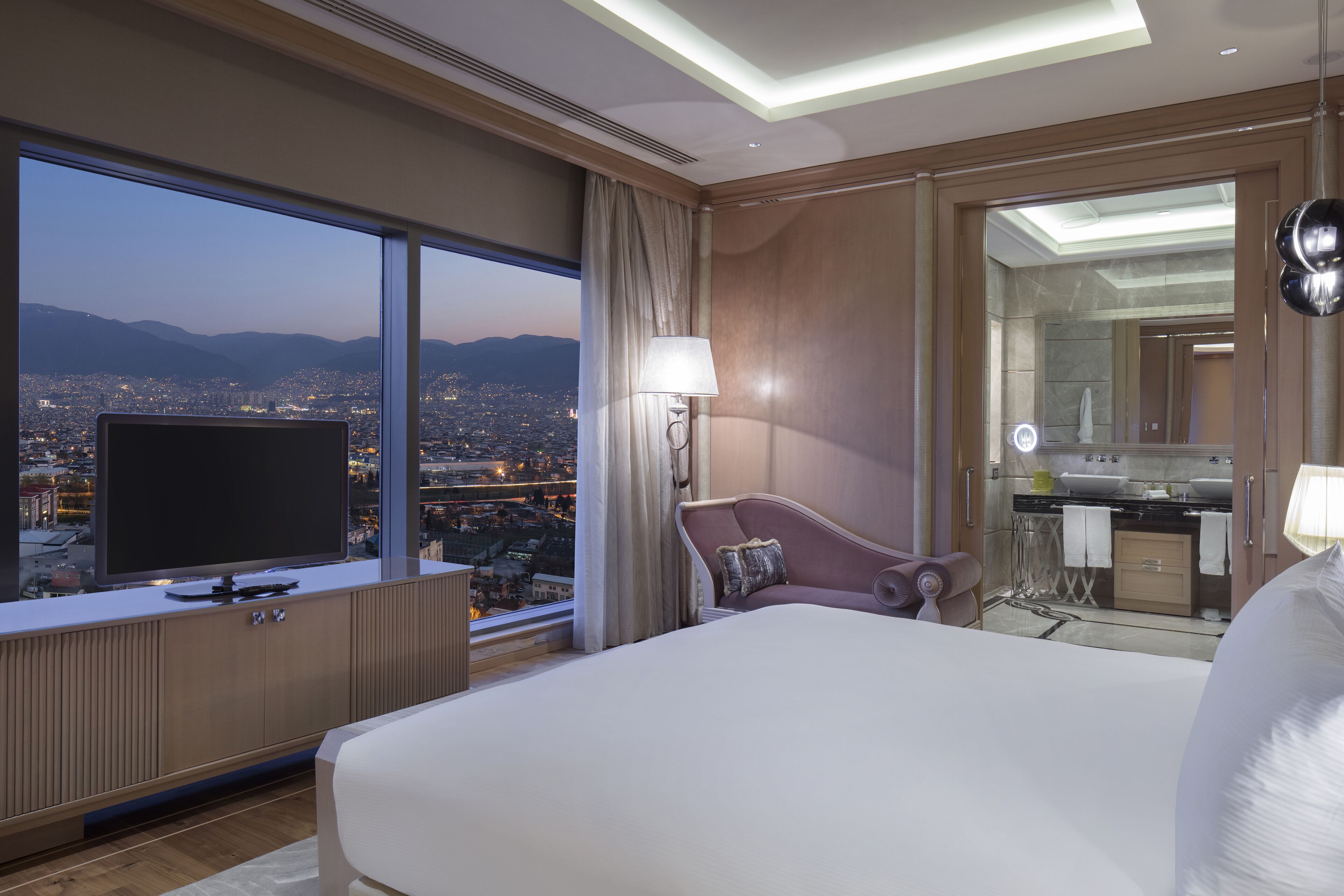 Une chambre d’hôtel avec un grand lit, un téléviseur posé sur une commode en bois, une chaise, un bureau avec un miroir et une vue sur le paysage urbain au crépuscule.