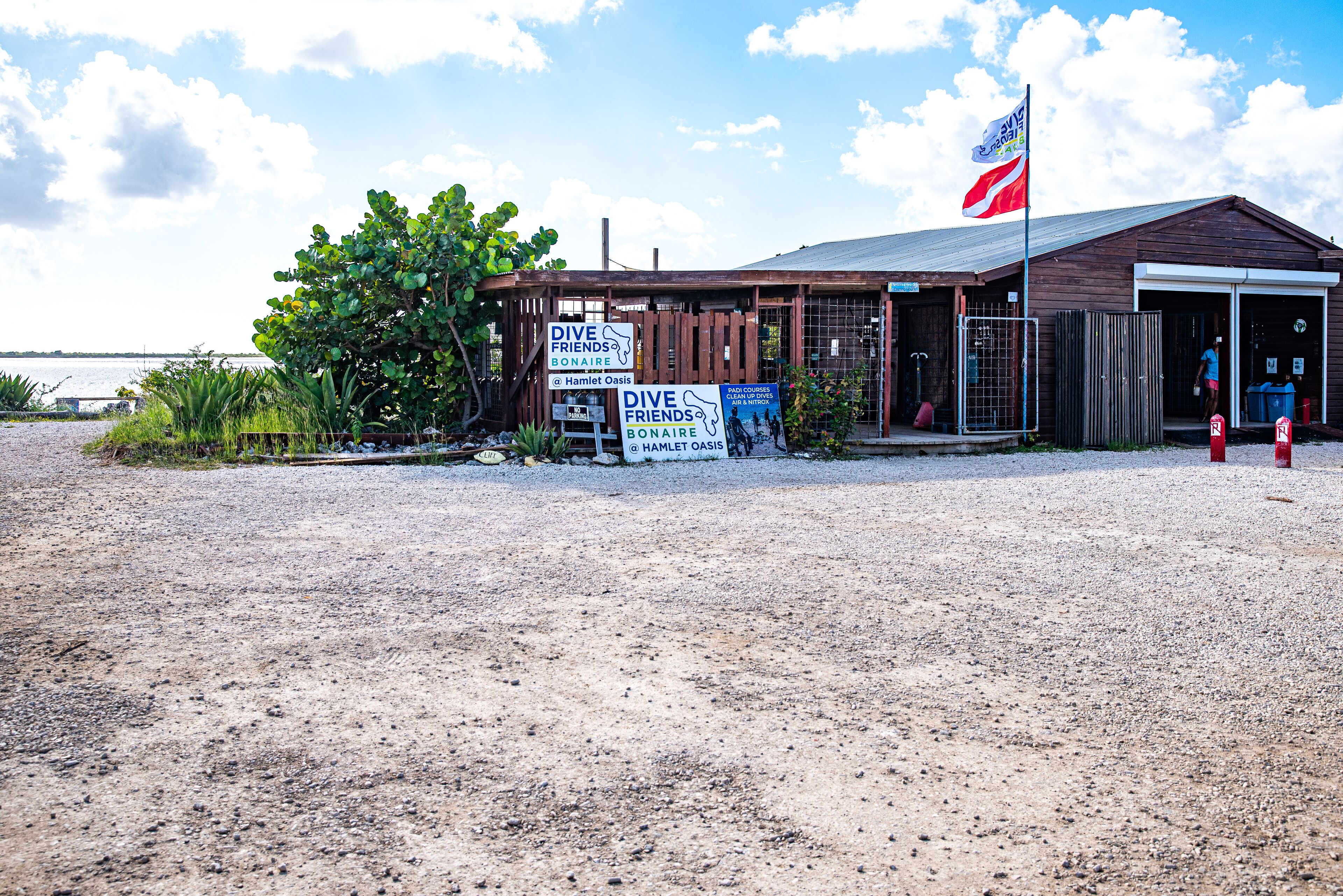 Een houten gebouw met een vlag, borden voor 'Dive Friends Bonaire' en 'Hamlet Oasis', en een grindpad ervoor.