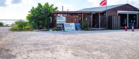 Een houten gebouw met een vlag, borden voor 'Dive Friends Bonaire' en 'Hamlet Oasis', en een grindpad ervoor.