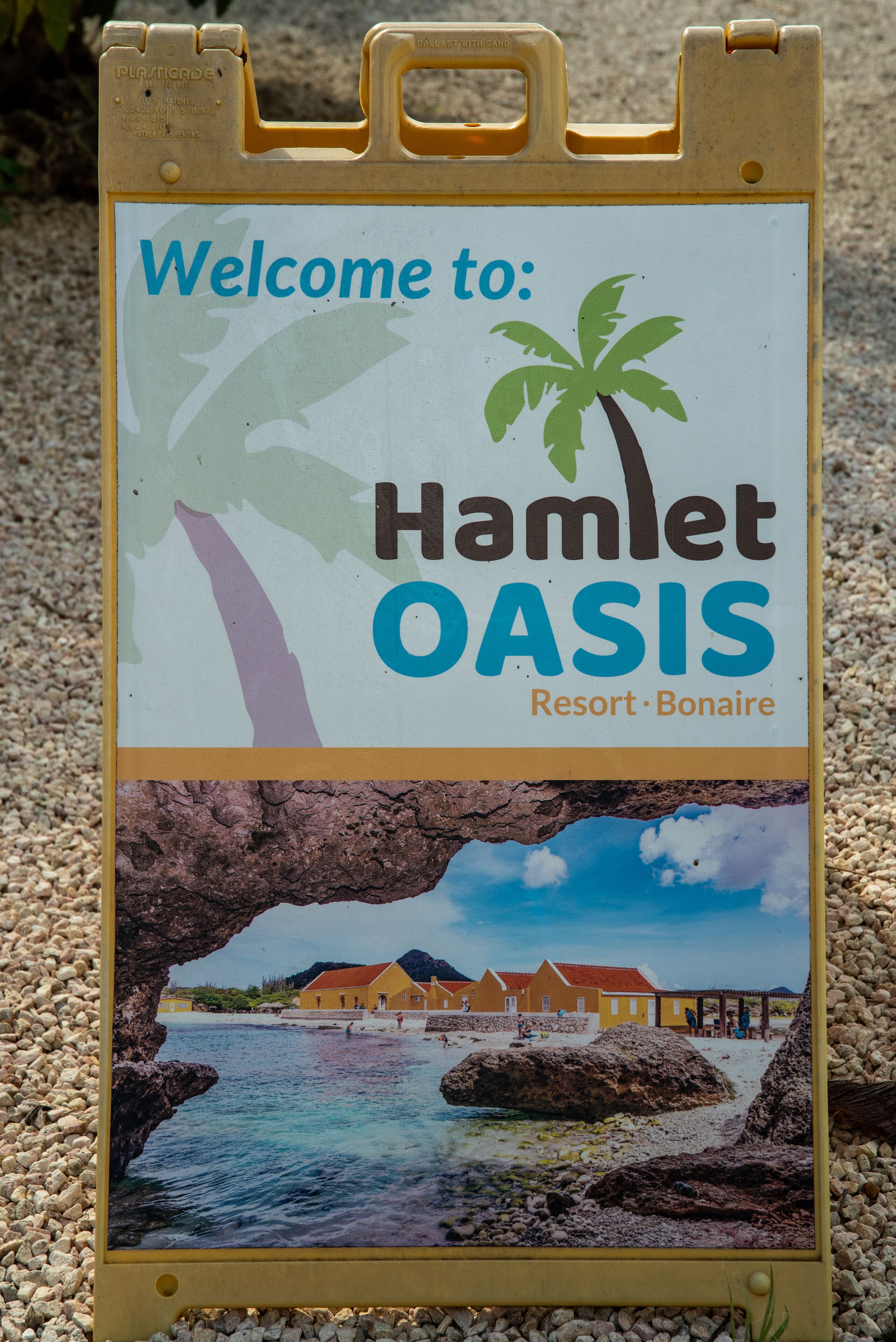 Segnaletica di benvenuto per Hamlet Oasis Resort, con una palma e una veduta costiera con edifici.