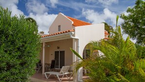 A white building with a red-tiled roof, a covered patio with white lounge chairs, and lush green foliage.