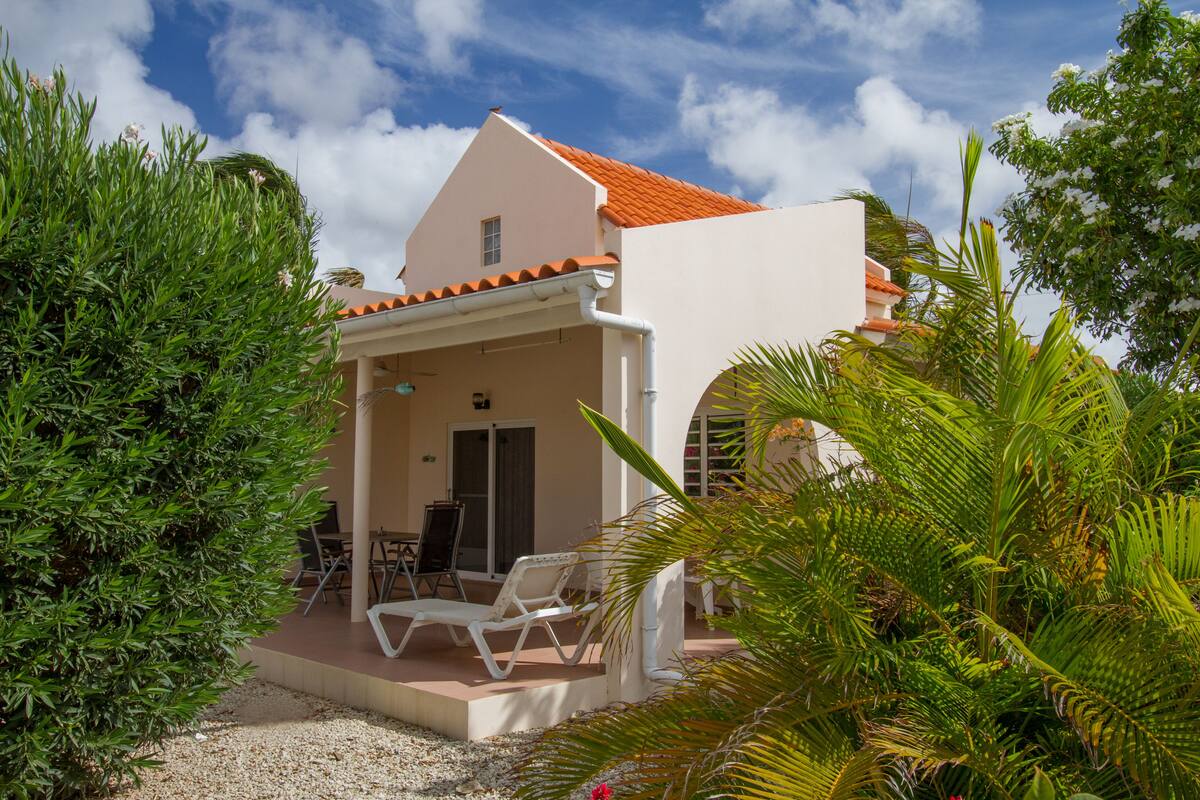 Un bâtiment blanc surmonté d’un toit de tuiles rouges, une terrasse couverte avec des chaises longues blanches et une végétation luxuriante.