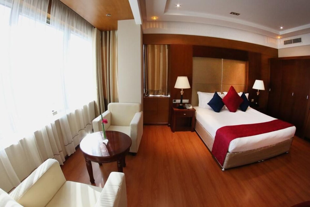 A hotel room with a bed, two bedside tables, a chair, a small table with a vase, and a window with curtains.