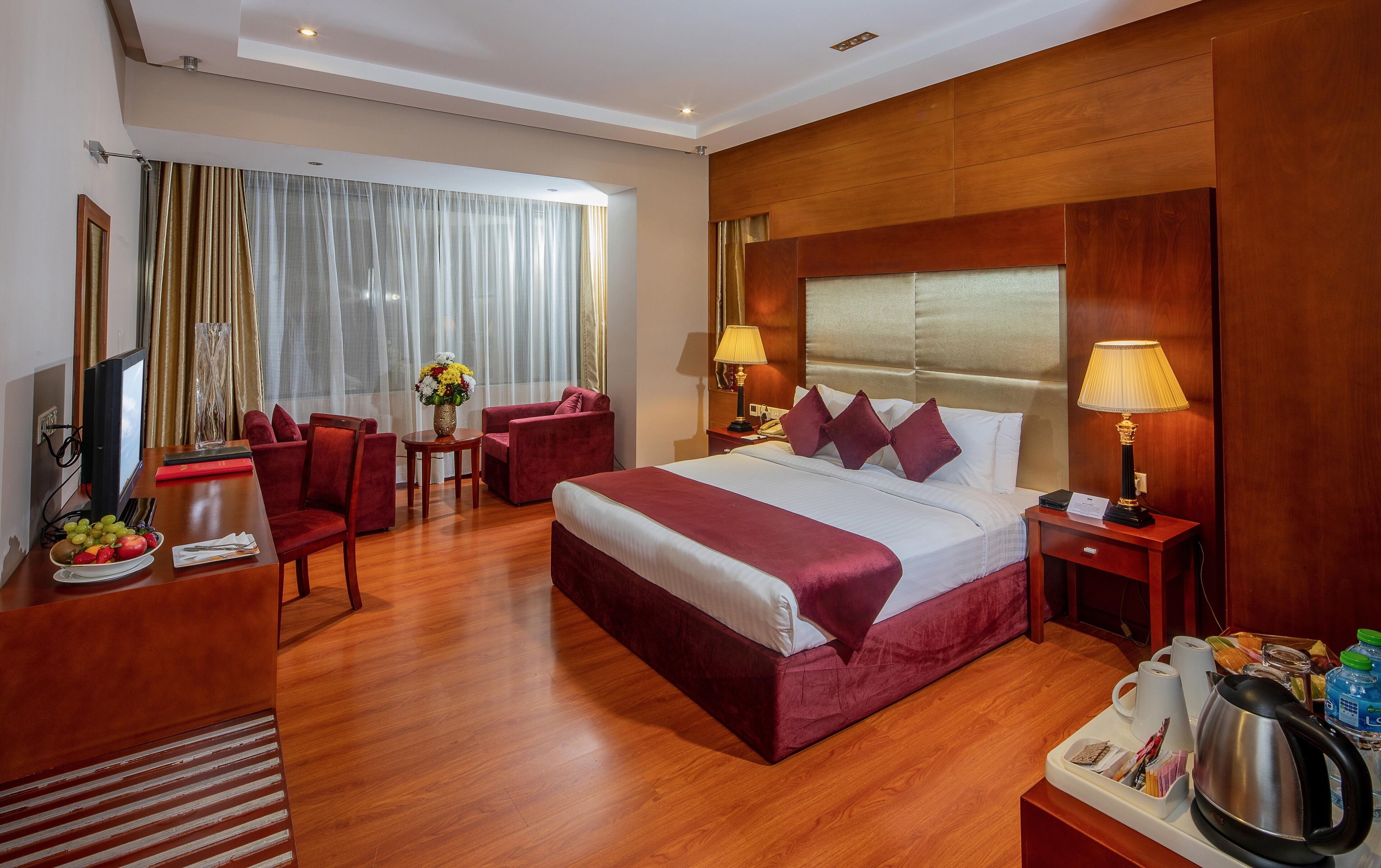 Una habitación de hotel con una cama grande, dos sillones rojos, un escritorio de madera con televisión y una mesa de comedor con fruta y un florero.