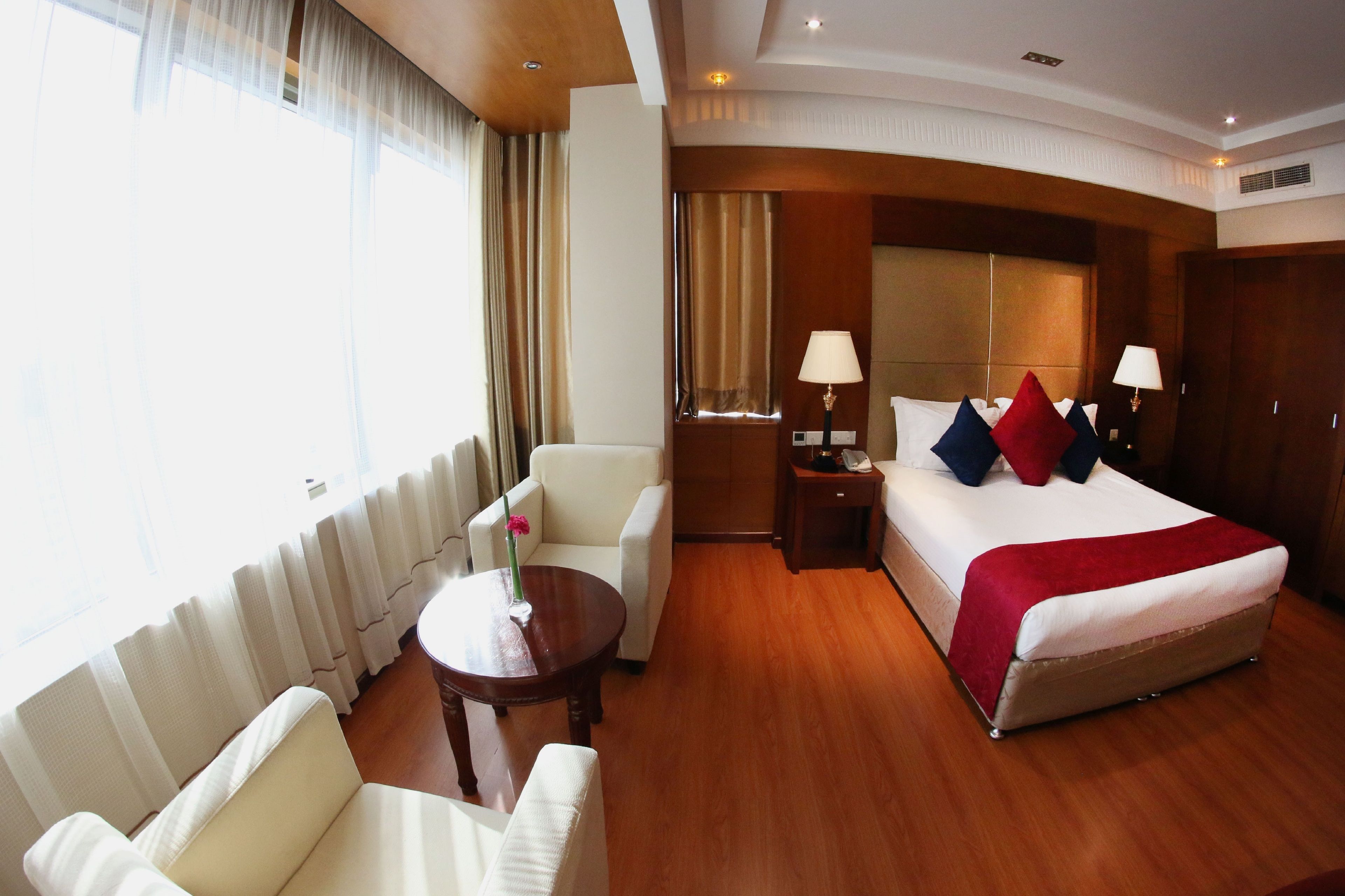 A hotel room with a bed, two bedside tables, a chair, a small table with a vase, and a large window with curtains.