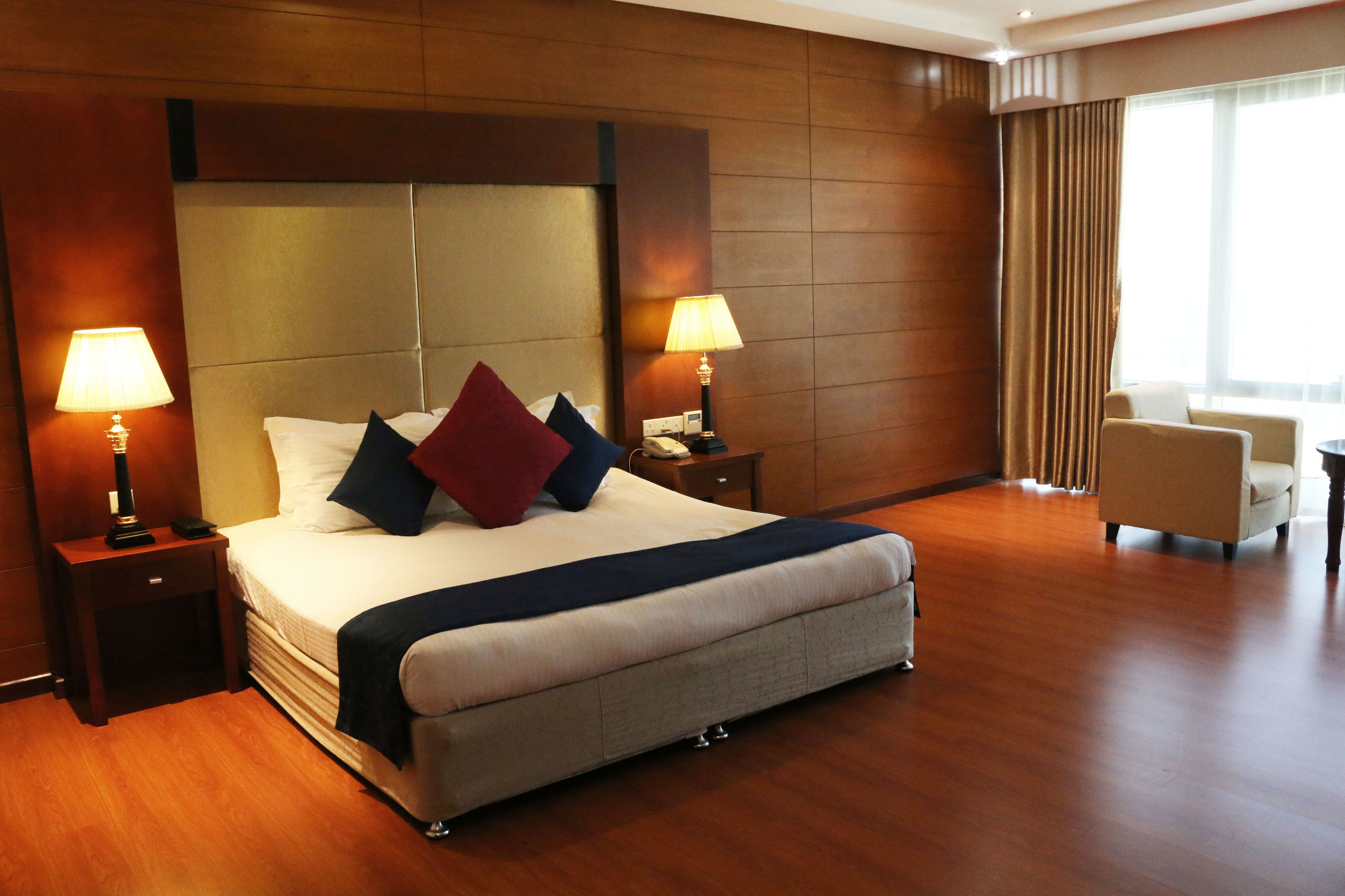 A hotel room with a large bed, two bedside tables with lamps, a chair, and a large window with curtains.