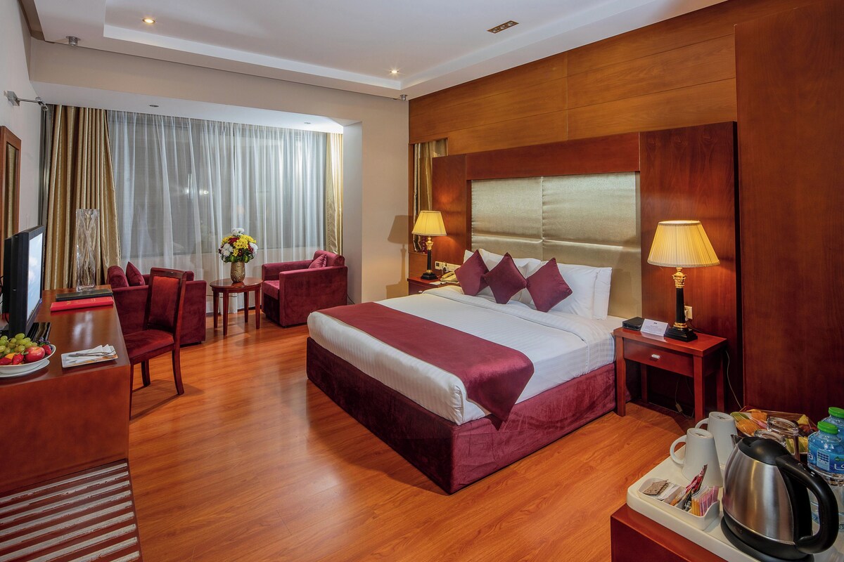 A hotel room with a large bed, two red armchairs, a wooden desk with a television, and a dining table with fruit and a vase of flowers.