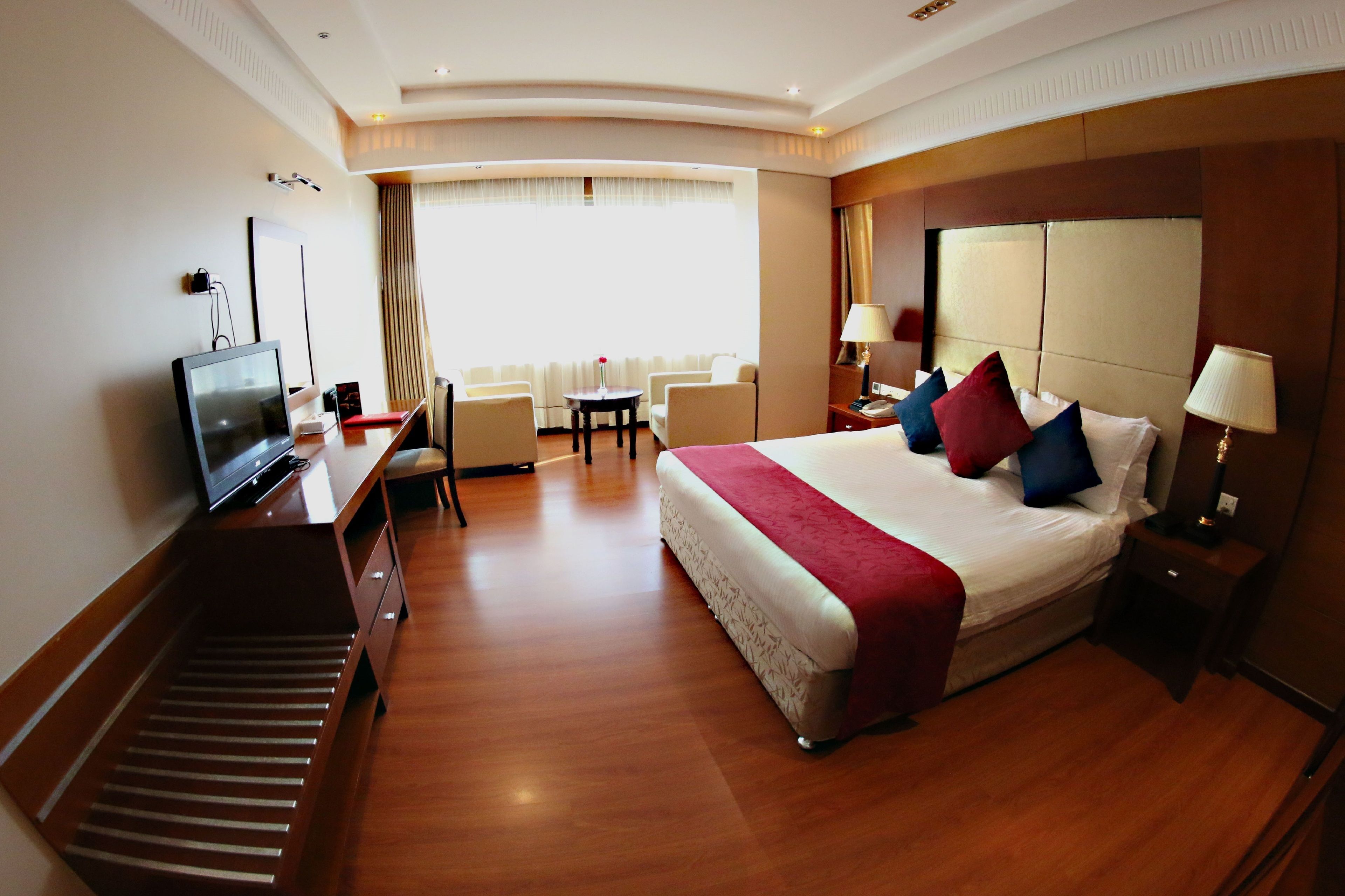 Habitación de hotel con cama, mesita de noche, lámpara, televisión, escritorio, silla y ventana con cortinas.