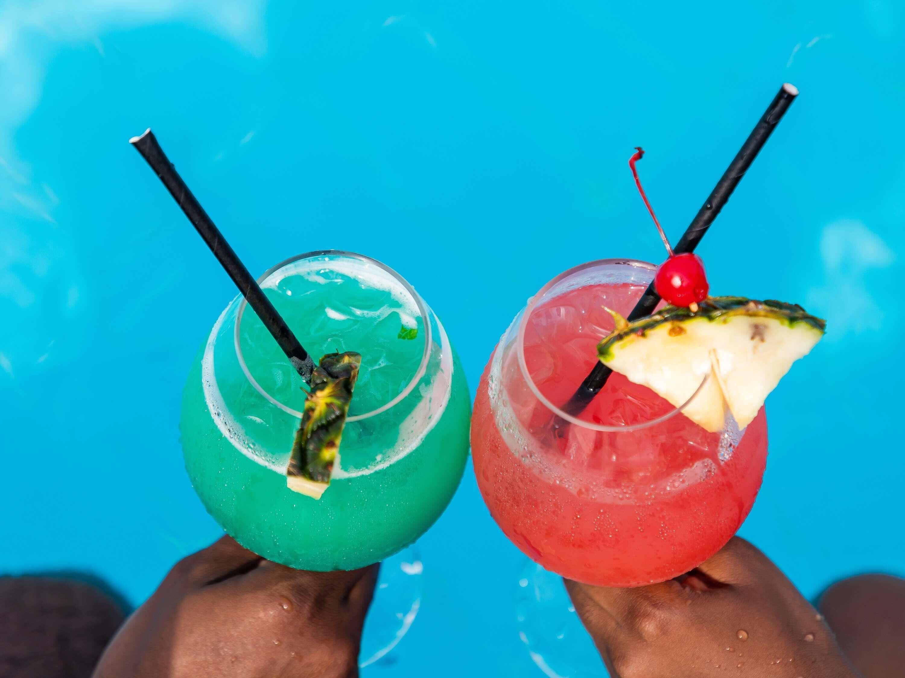 Dos bebidas con pajitas y adornos, sostenidas por manos sobre un fondo azul.