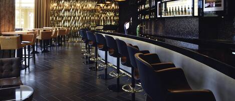 Zona de bar con taburetes altos, una barra y estantes con botellas.
