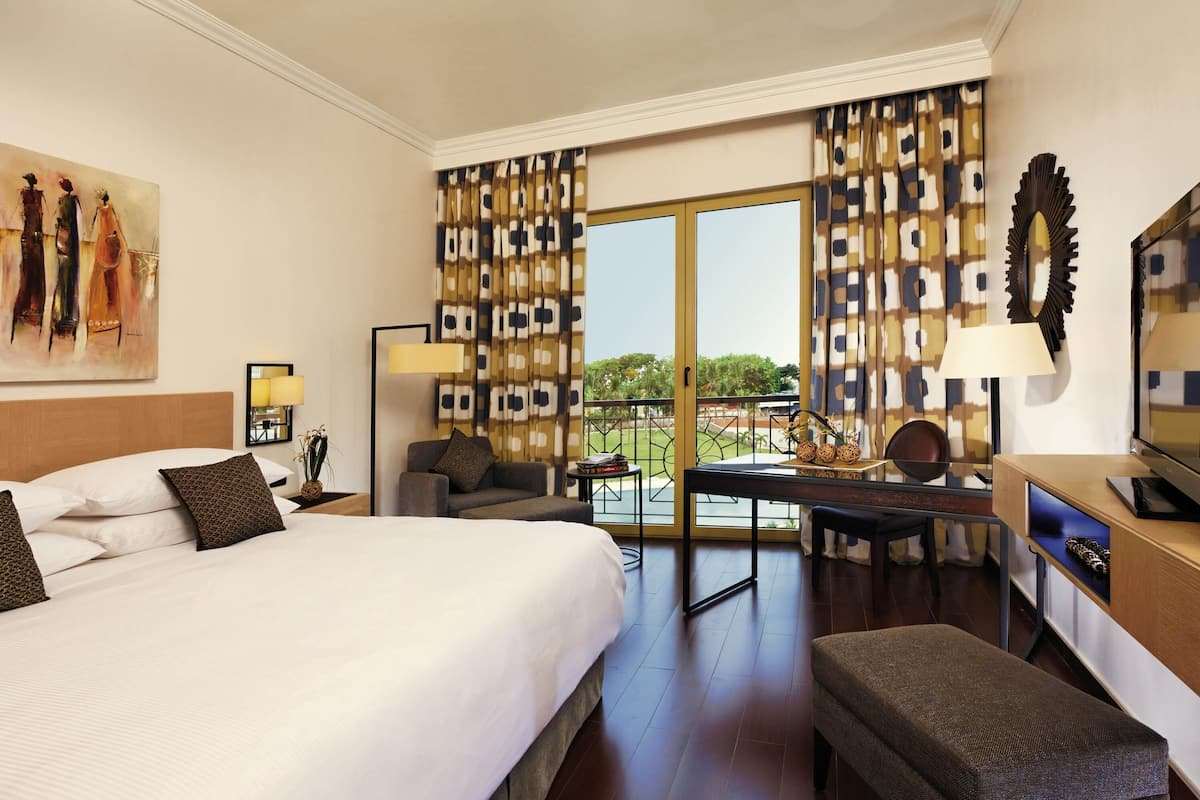 Ein Hotelzimmer mit einem großen Bett, einem Schreibtisch mit Fernseher, einem Sessel und einem Balkon mit gemusterten Vorhängen.