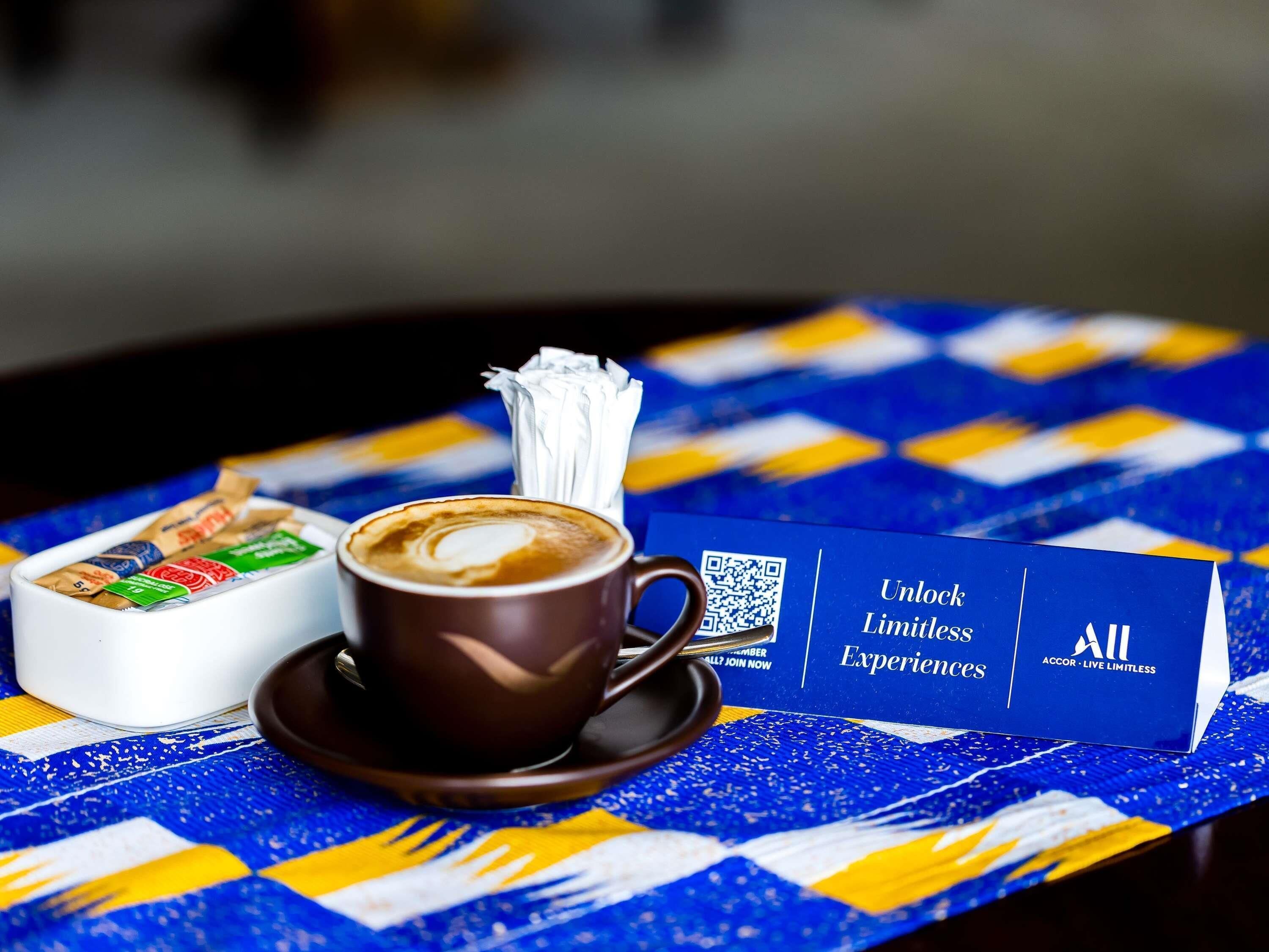 Una taza de café con arte de latte, una servilleta y un código QR sobre una mesa con mantel de estampado azul y amarillo.