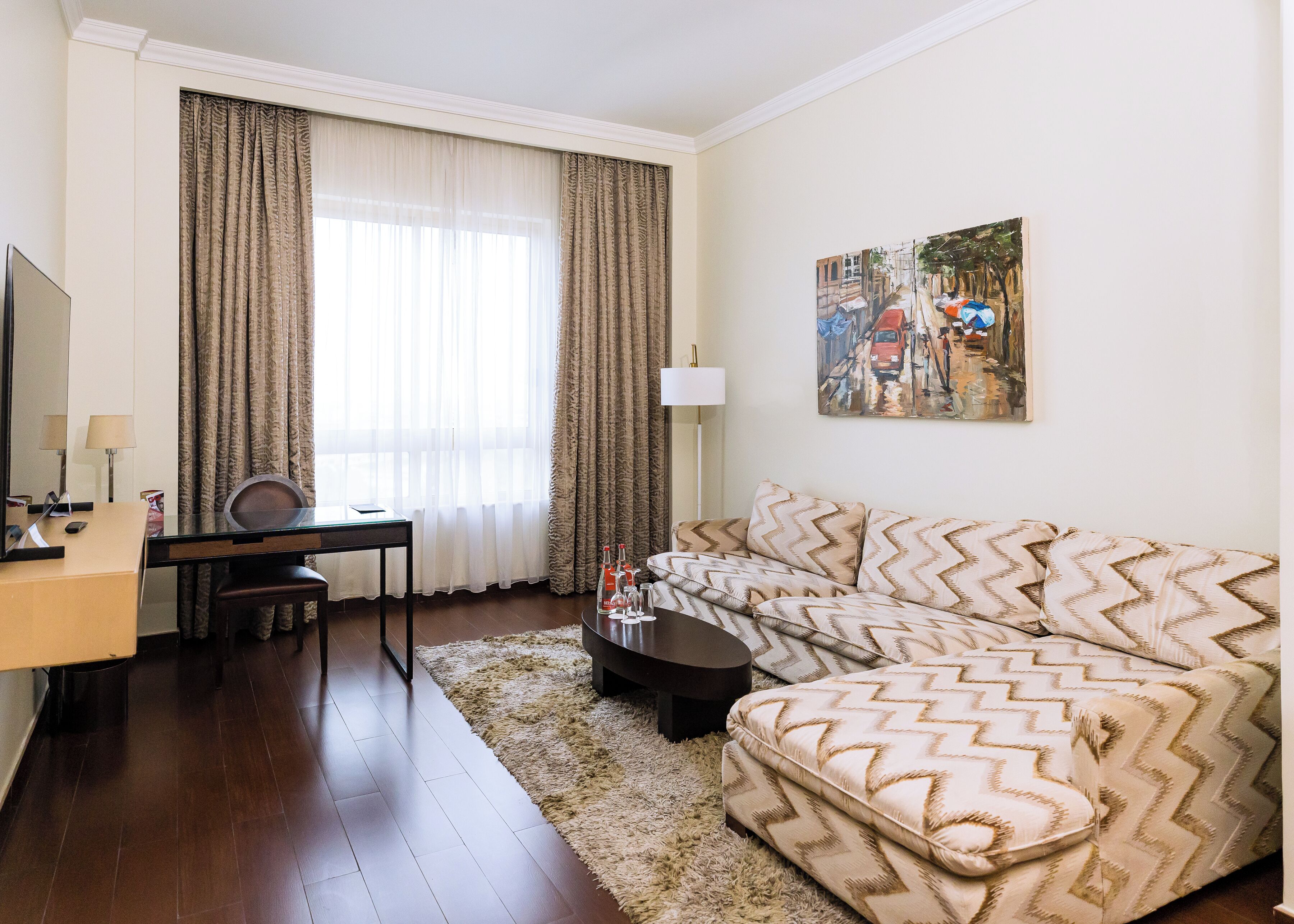 A hotel room with a sofa, a desk, a chair, a lamp, and a painting on the wall.