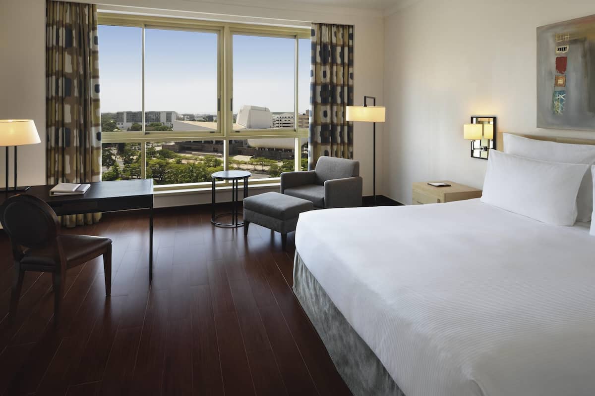 Ein Hotelzimmer mit einem großen Bett, einem Schreibtisch mit Stuhl, einem kleinen Tisch und Blick auf Gebäude und Bäume durch das Fenster.