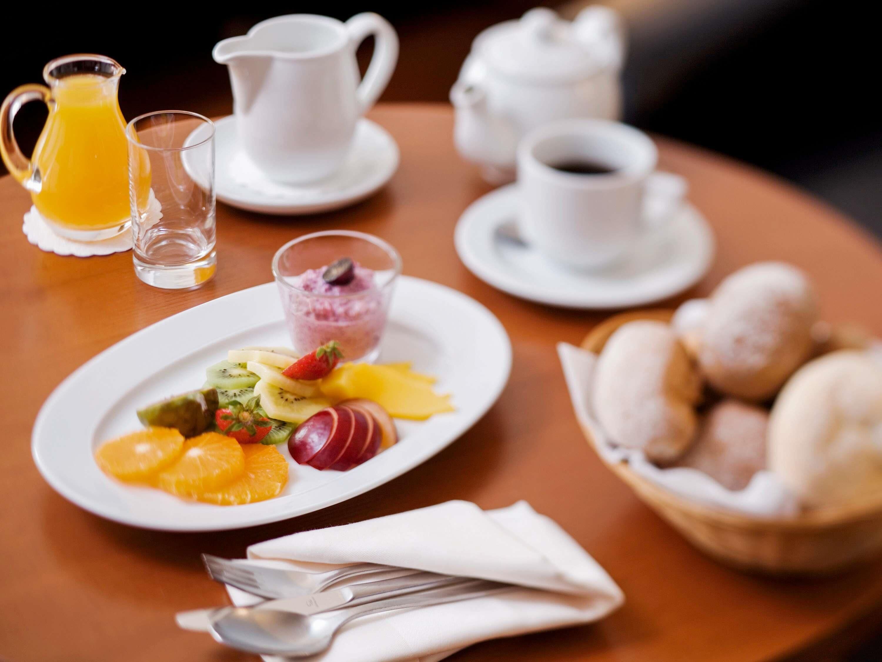 Une table mise pour le petit-déjeuner avec une assiette de fruits, un pichet de jus d’orange, une tasse de café et un bol de viennoiseries.