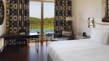 Quarto de hotel com cama, escrivaninha, cadeira, sofá e vista para a piscina e vegetação.