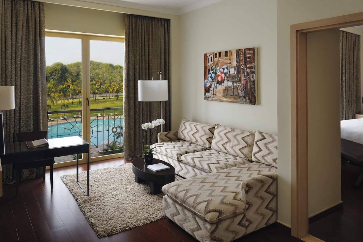 Ein Hotelzimmer mit einer Couch, einem Schreibtisch, einer Lampe, einem Gemälde und Blick auf einen Pool.