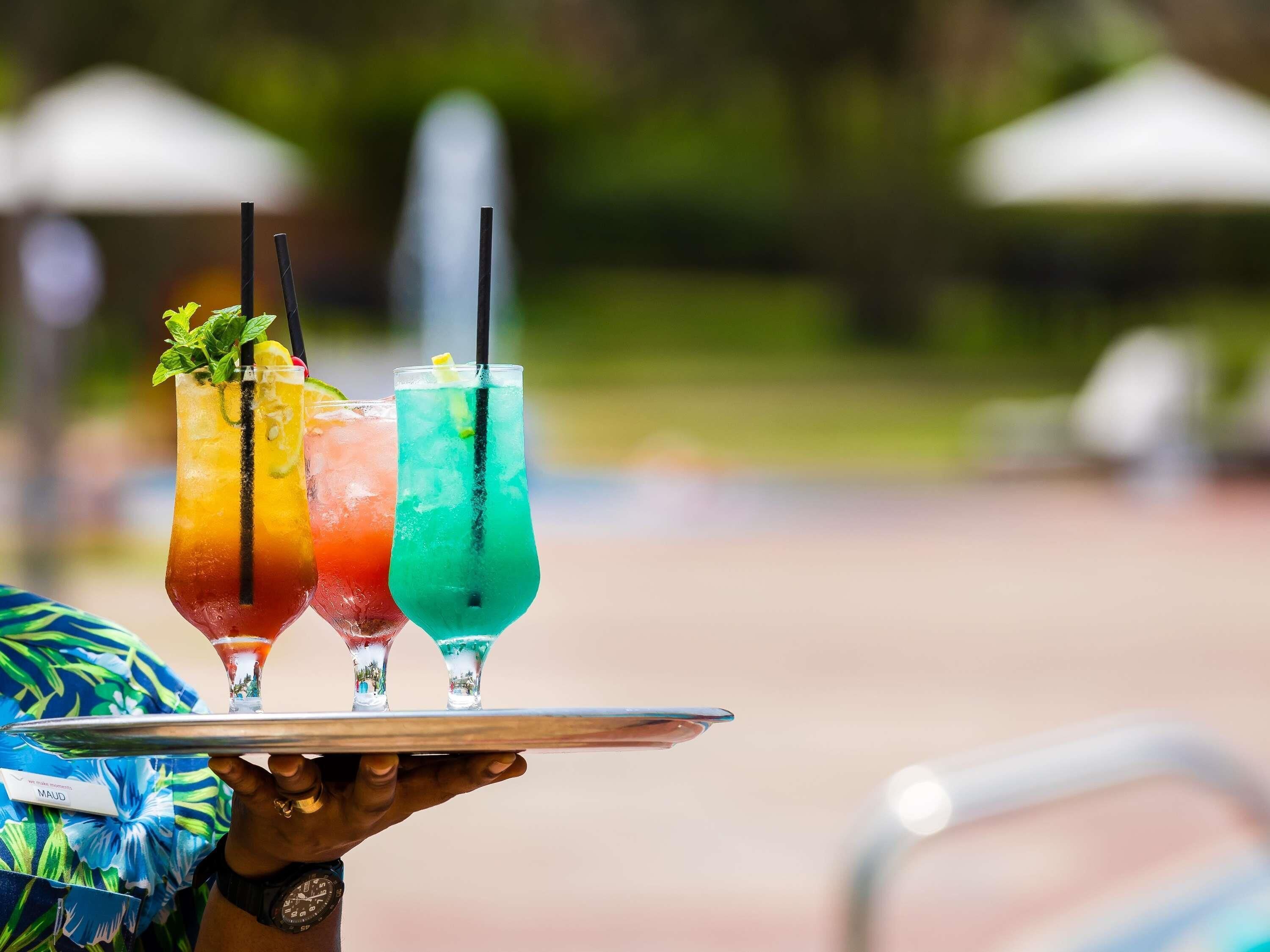 A person holding a tray with three colorful drinks, each with a straw and garnish, outdoors.
