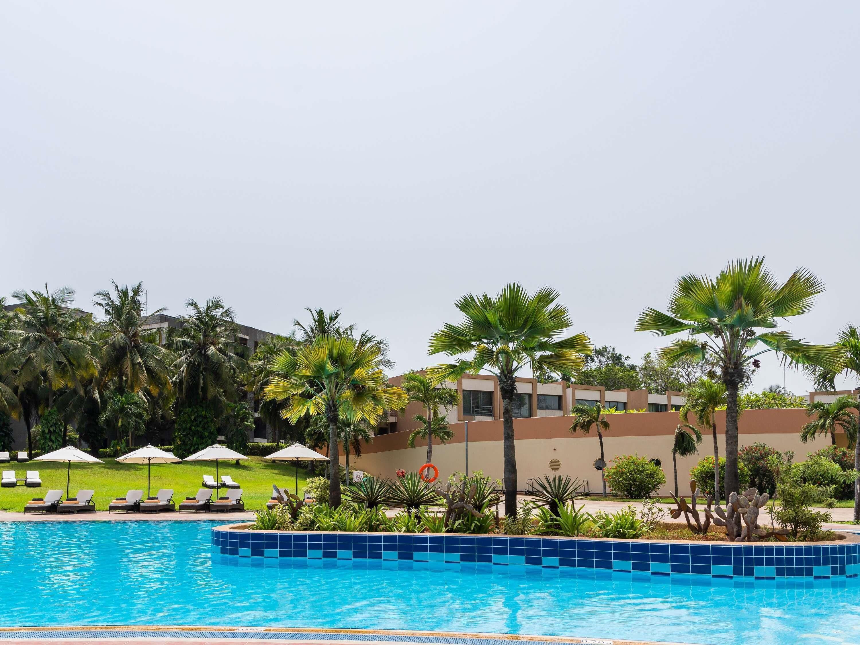 A swimming pool with lounge chairs and umbrellas, surrounded by palm trees and a building with a beige facade.