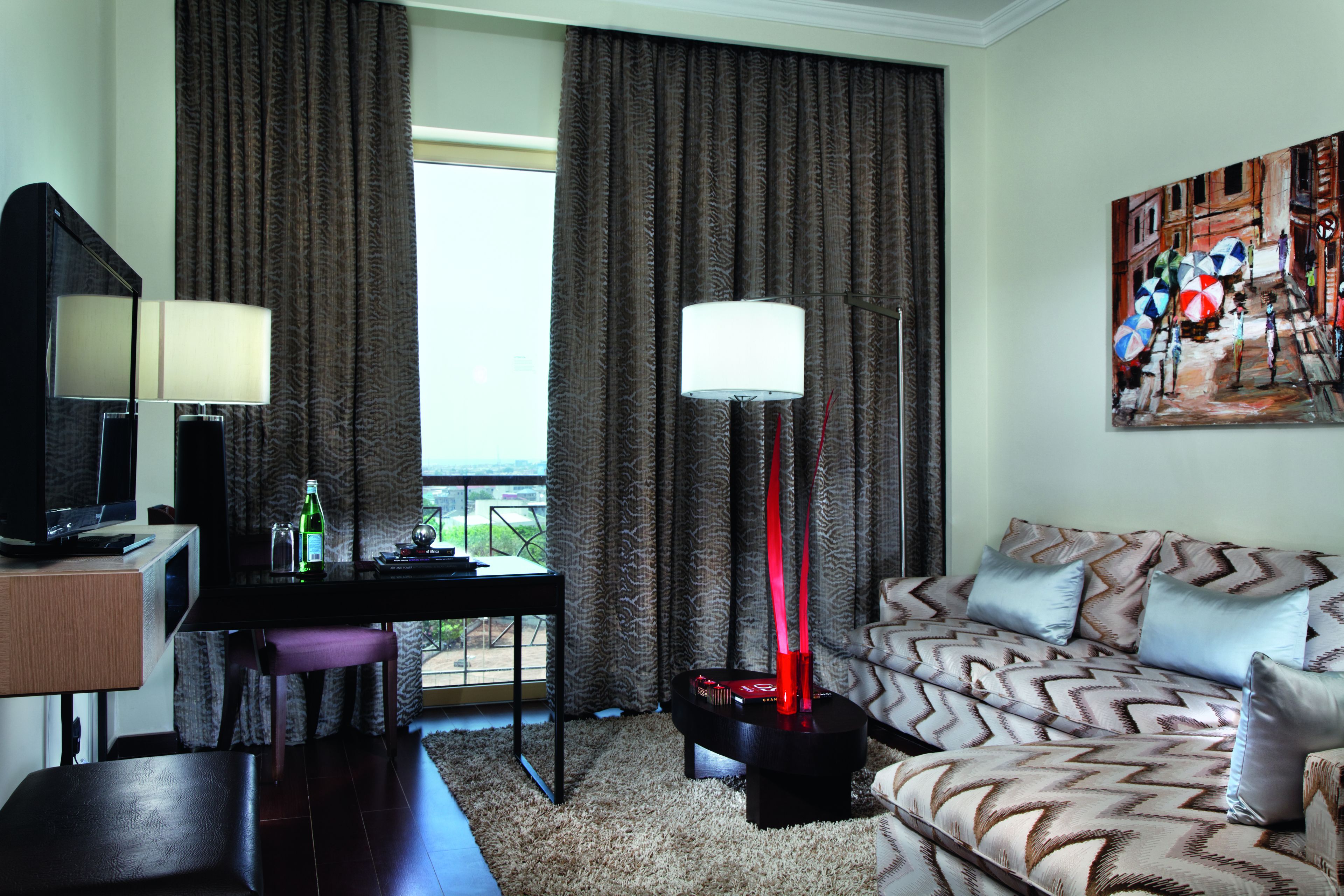 A hotel room with a sofa, a TV, a desk with a lamp, a painting on the wall, and a view of the outdoors.