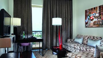 A hotel room with a sofa, a TV, a desk with a lamp, a painting on the wall, and a view of the outdoors.