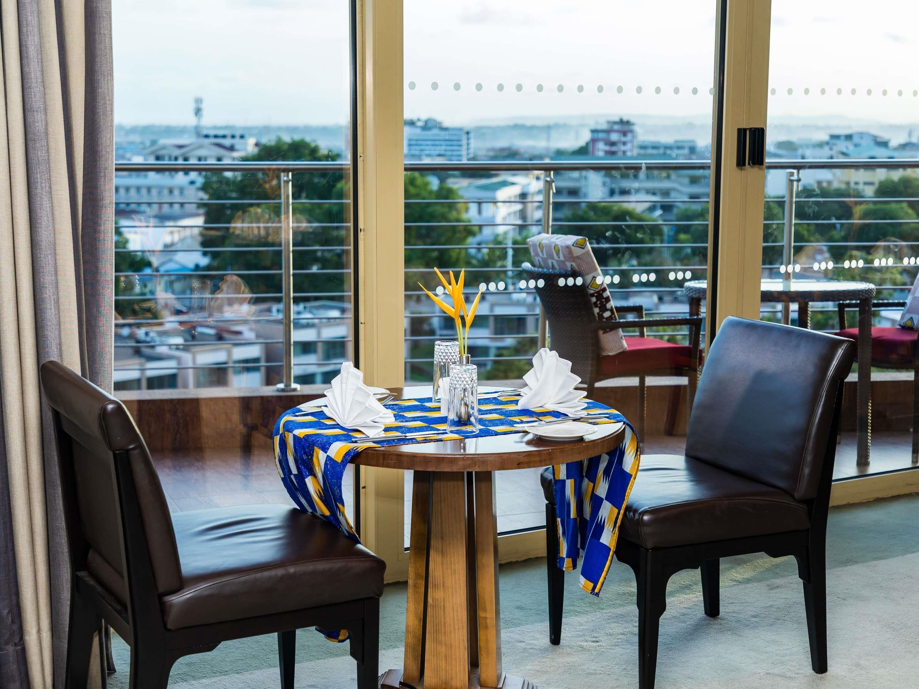 A dining area with a table set for two, a view of the cityscape through large windows, and a balcony railing.