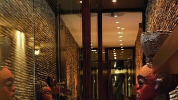 A corridor with a patterned carpet, glass walls reflecting statues, and a wooden bench.