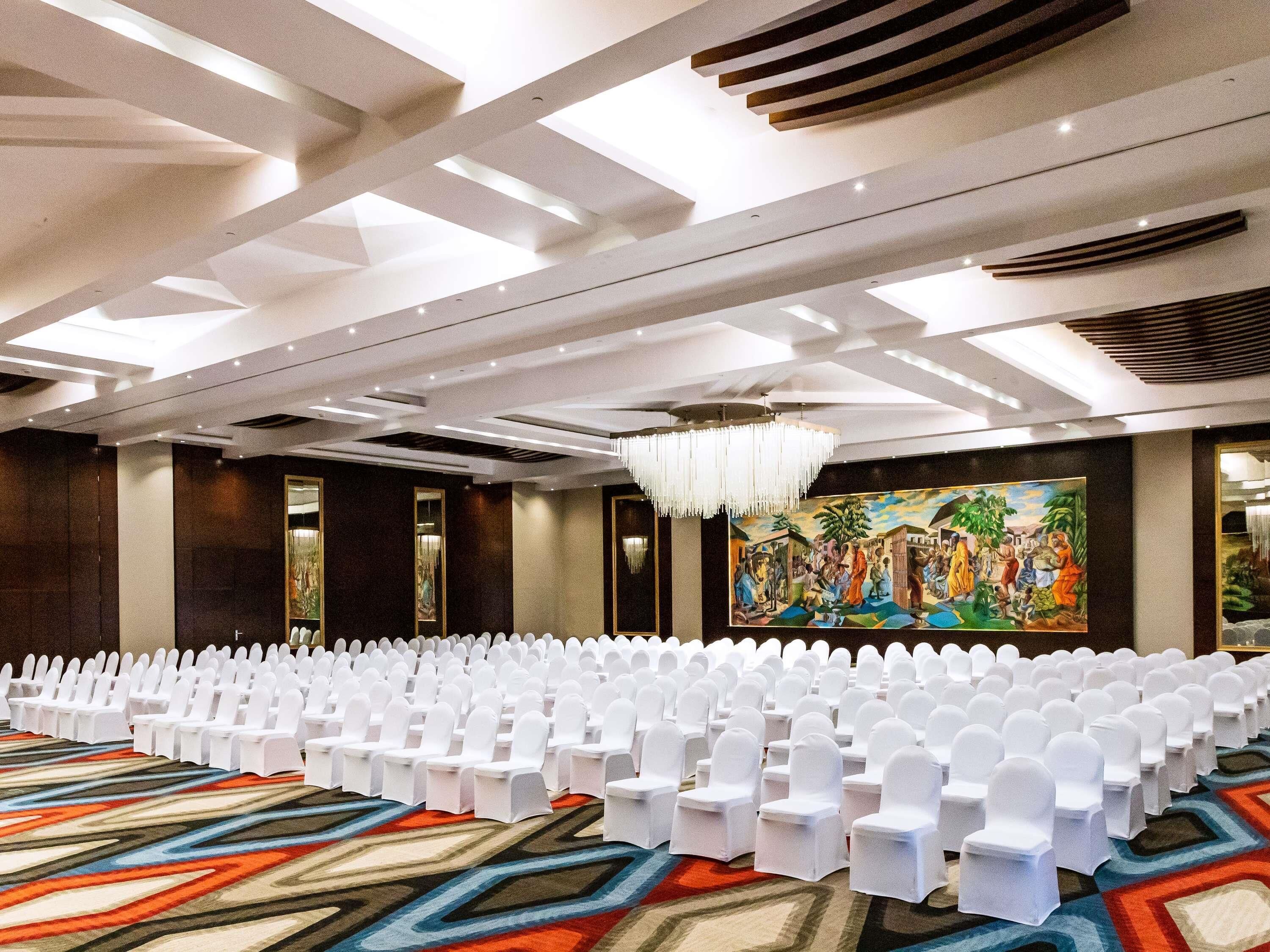 A conference room with rows of white chairs, a colorful carpet, and a large painting on the wall.