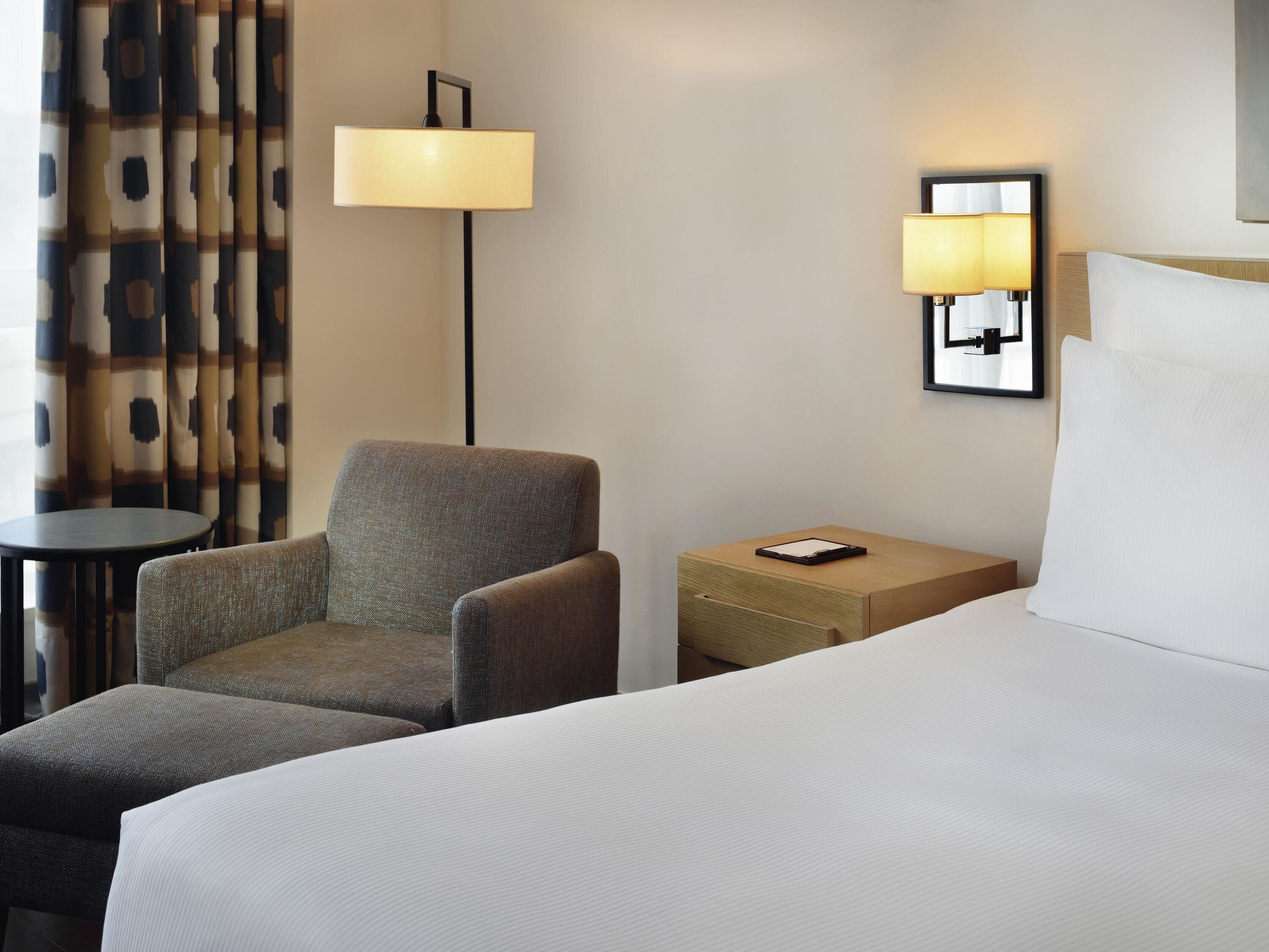 Habitación de hotel con una cama, una silla, una mesita pequeña y dos lámparas fijadas a la pared.