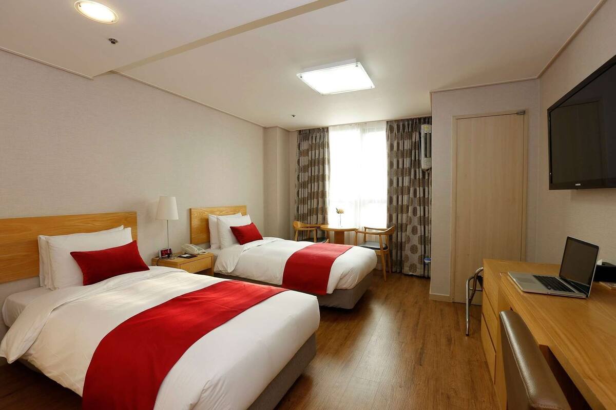 Una habitación de hotel con dos camas, un escritorio, una silla, un televisor y un ventanal con cortinas.