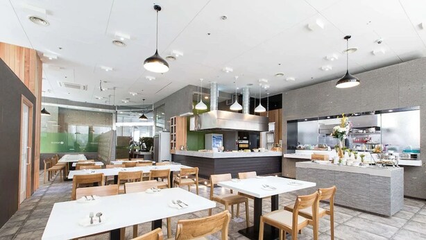 Un restaurant moderne avec des tables et des chaises blanches, un espace cuisine et des luminaires suspendus.