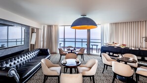 A hotel room with a leather sofa, dining area with chairs and tables, a large window with a view of the sea, and a buffet table with food and drinks.
