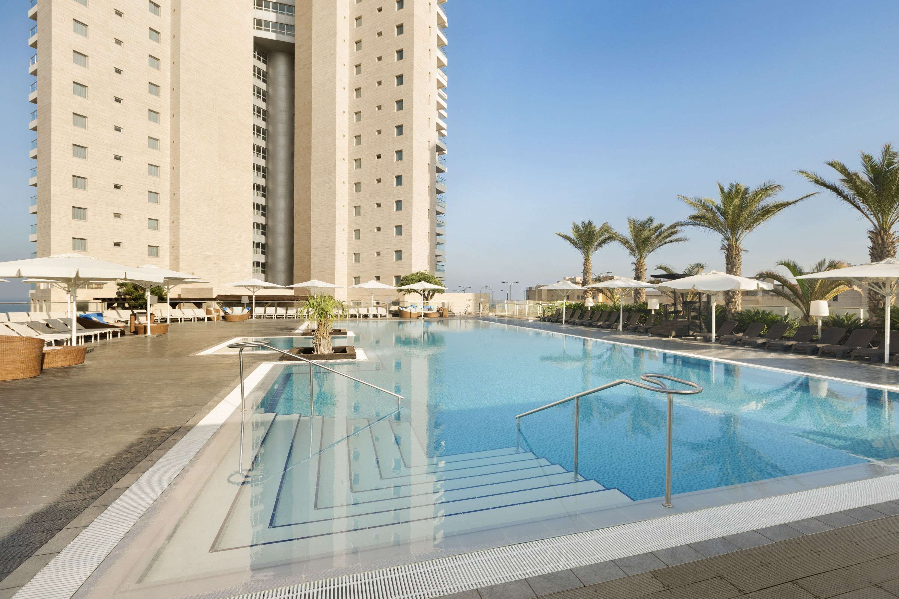 A swimming pool with lounge chairs and umbrellas, surrounded by palm trees and a tall building in the background.