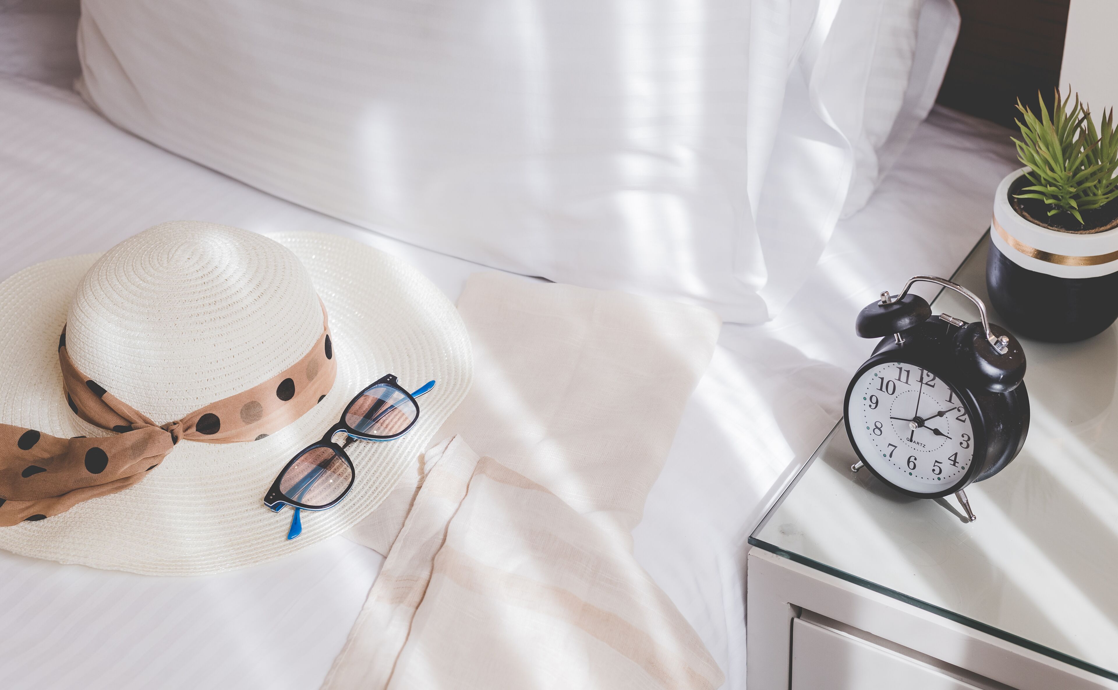 Una cama con una colcha blanca, un sombrero blanco con lazo marrón, un par de gafas de sol y un despertador negro sobre una mesita de noche.