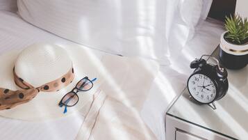 Una cama con una colcha blanca, un sombrero blanco con lazo marrón, un par de gafas de sol y un despertador negro sobre una mesita de noche.