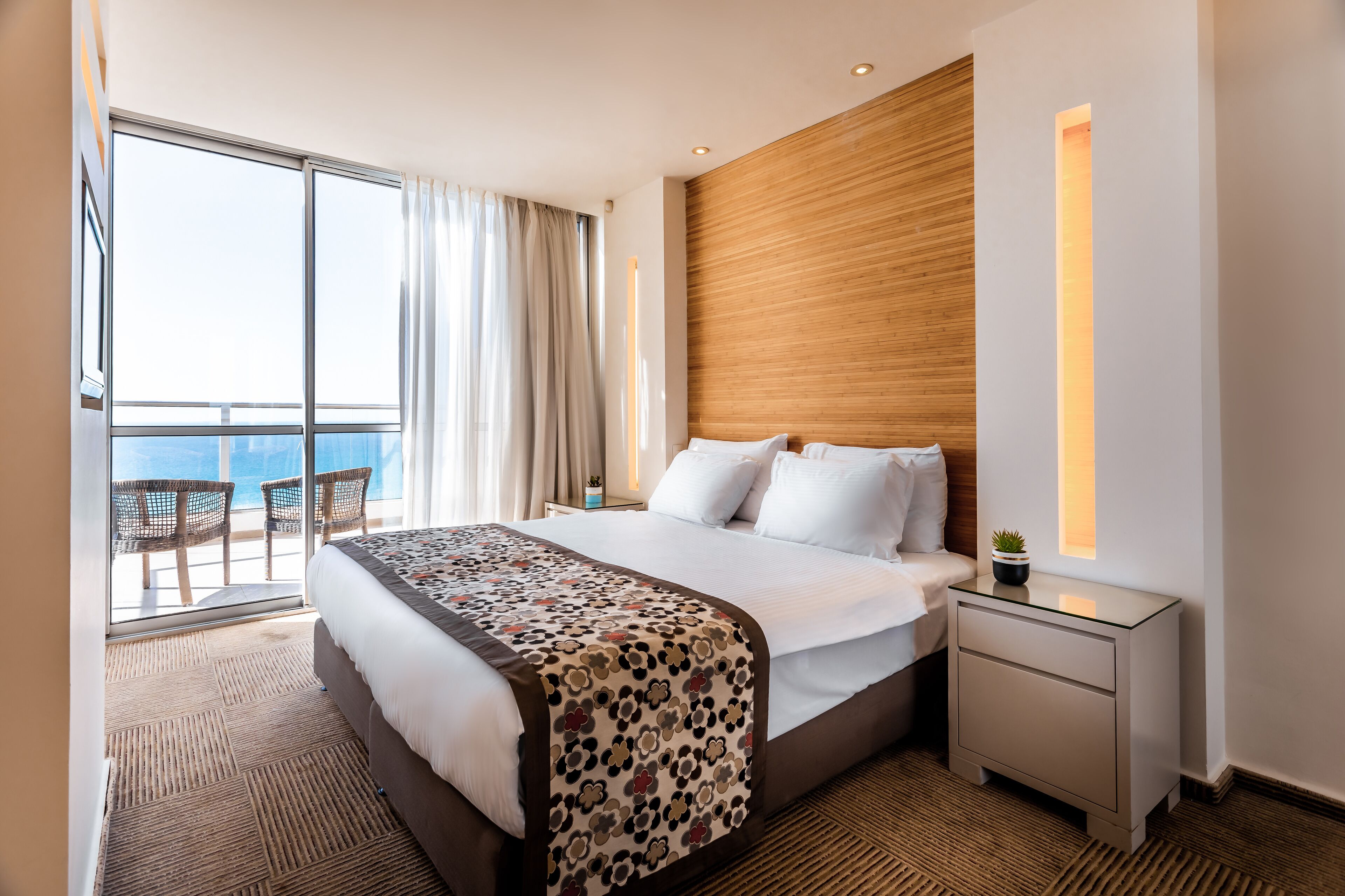 A hotel room with a large bed, a nightstand, a window with curtains, and a view of the sea.