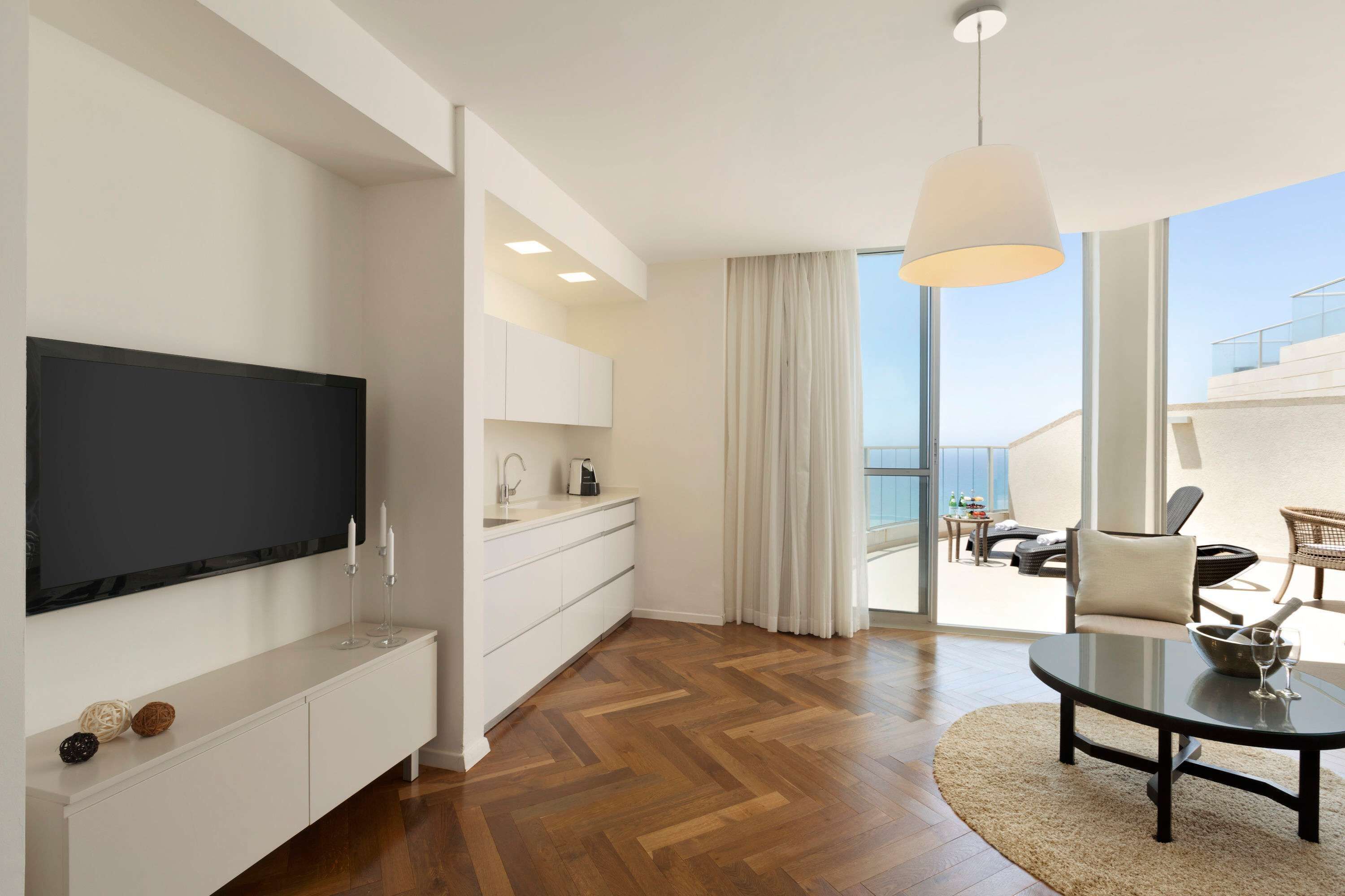 A modern living room with a flat-screen TV, a white console, a round coffee table, and a view of the balcony.