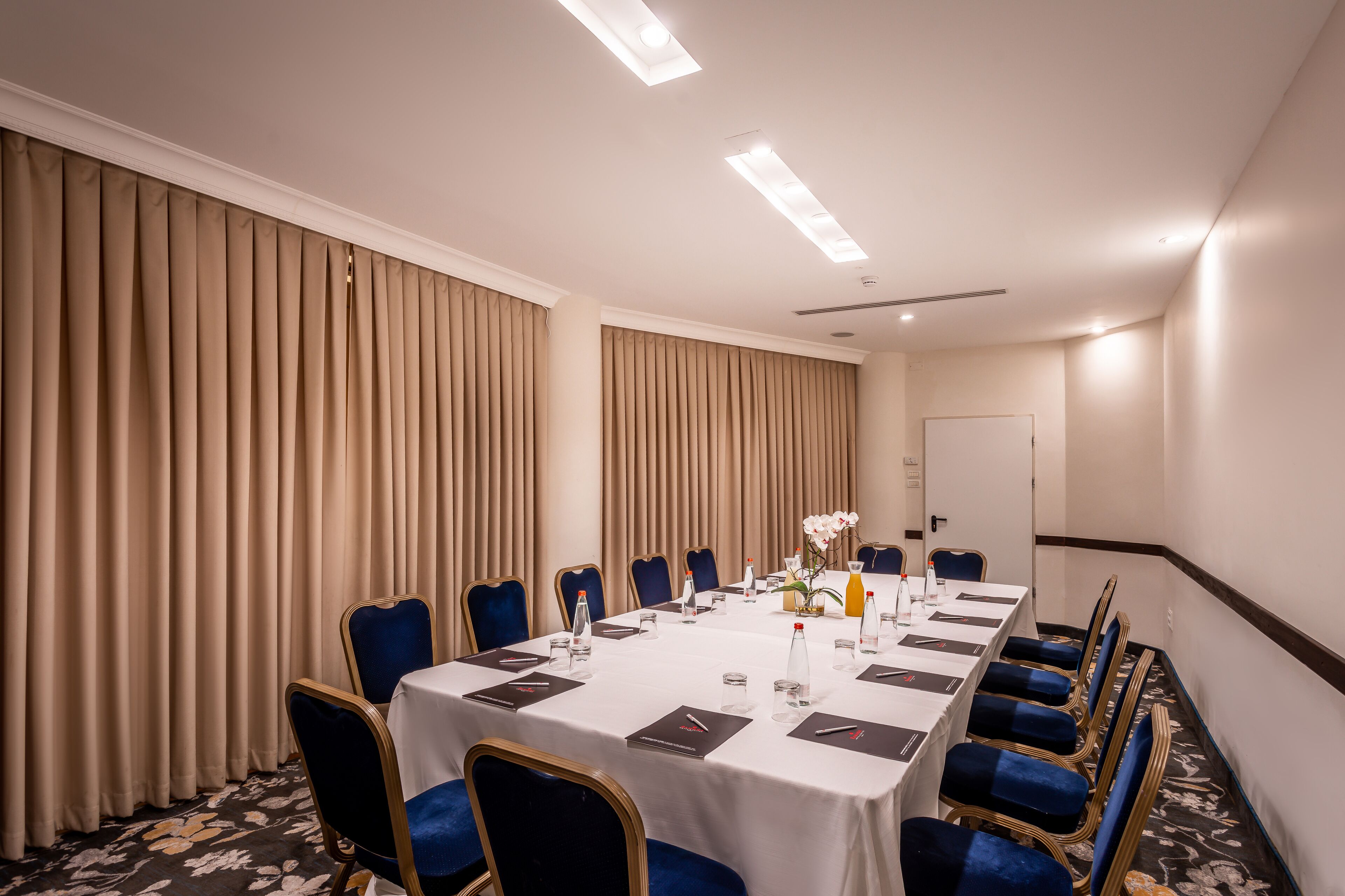 Ein Konferenzraum mit einem langen Tisch, blauen Stühlen und beigen Vorhängen.