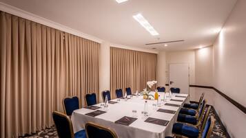 Ein Konferenzraum mit einem langen Tisch, blauen Stühlen und beigen Vorhängen.