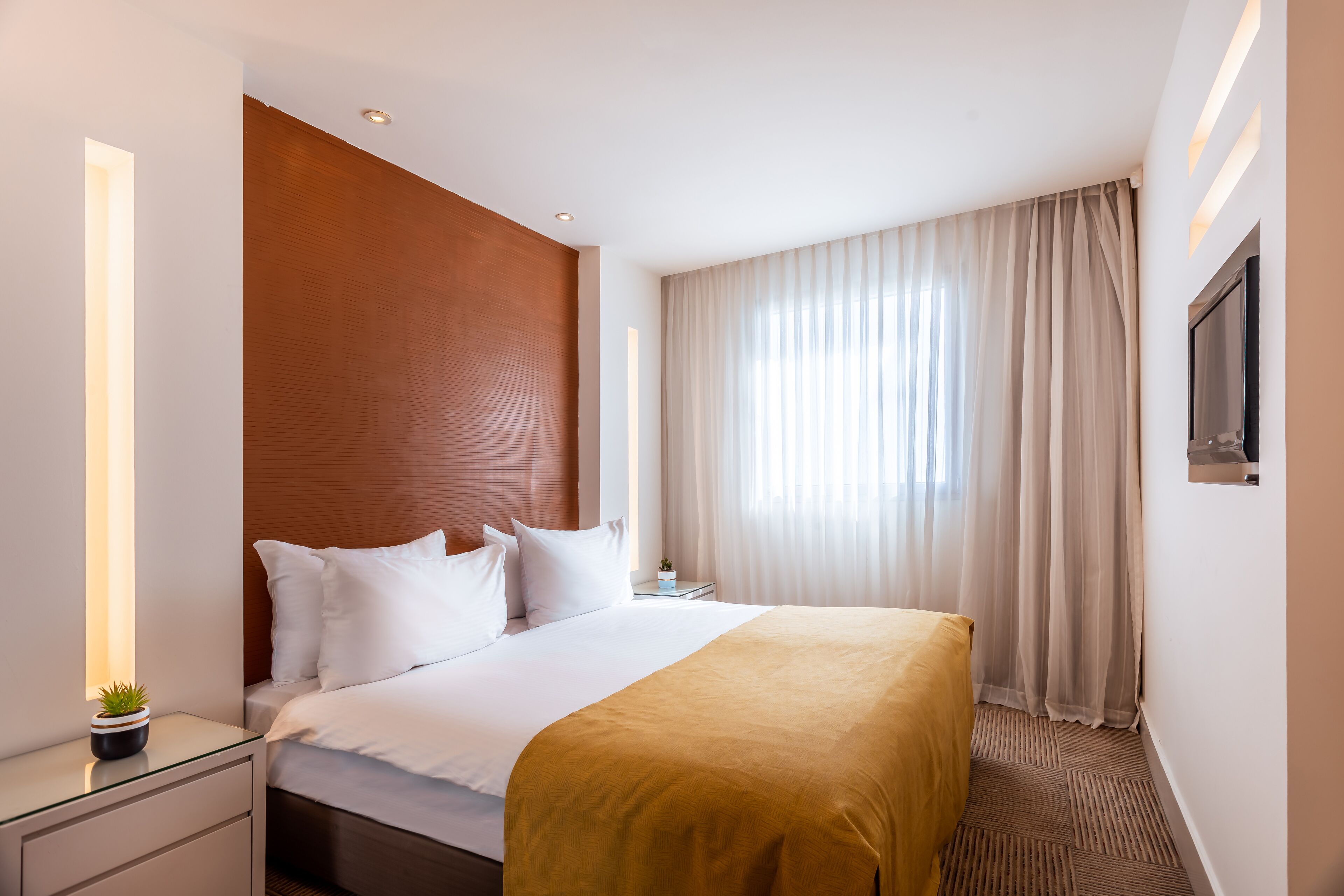 A modern hotel room with a large bed, a bedside table, a nightstand, a TV mounted on the wall, and a window with sheer curtains.