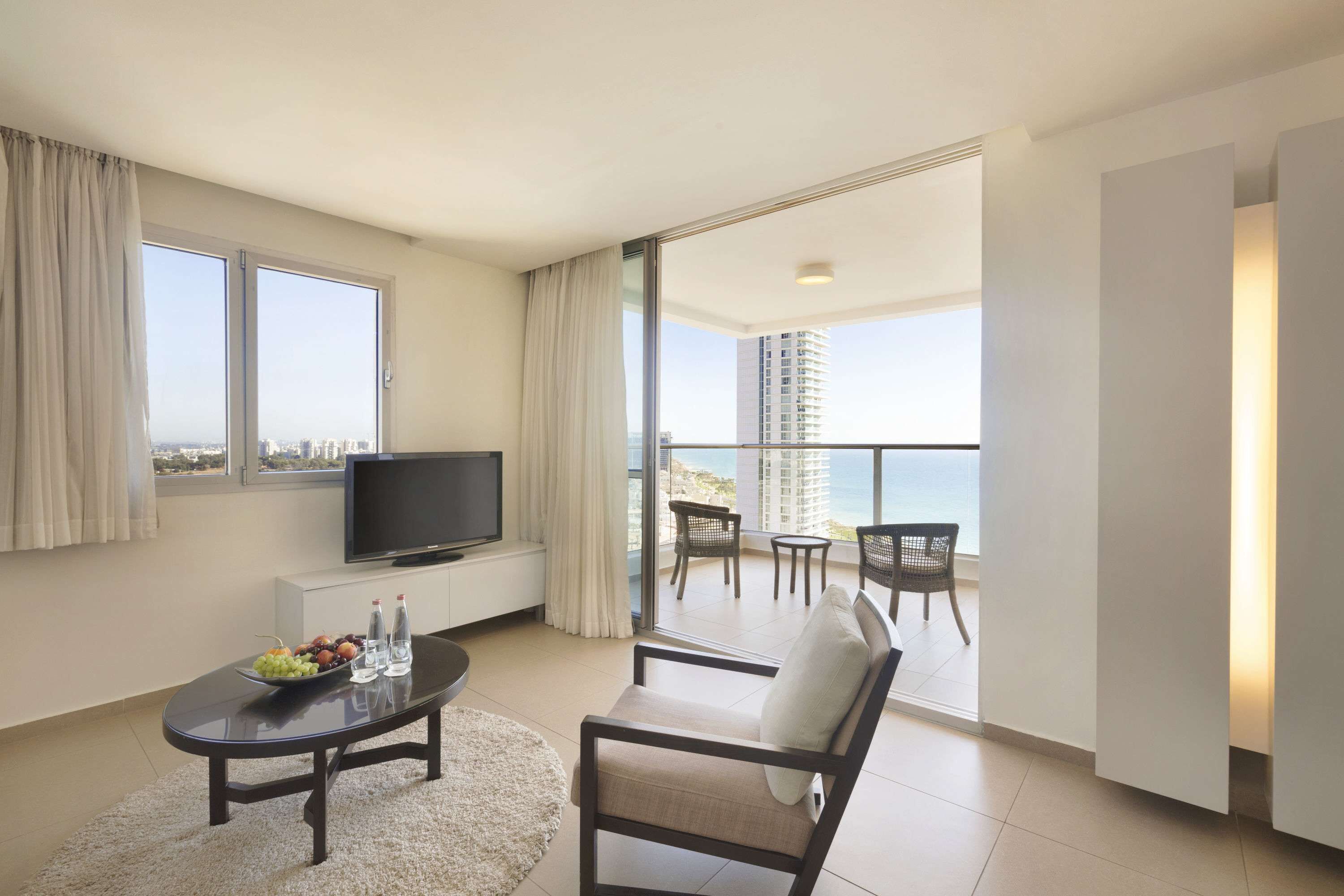 Habitación de hotel moderna con balcón, televisor de pantalla plana, una mesa de comedor con fruta y botellas de agua, y vistas a la ciudad y al océano.