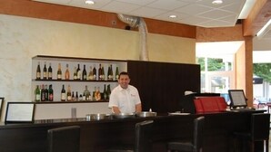 Una zona de bar con un bartender, una estantería de vinos y taburetes de bar.